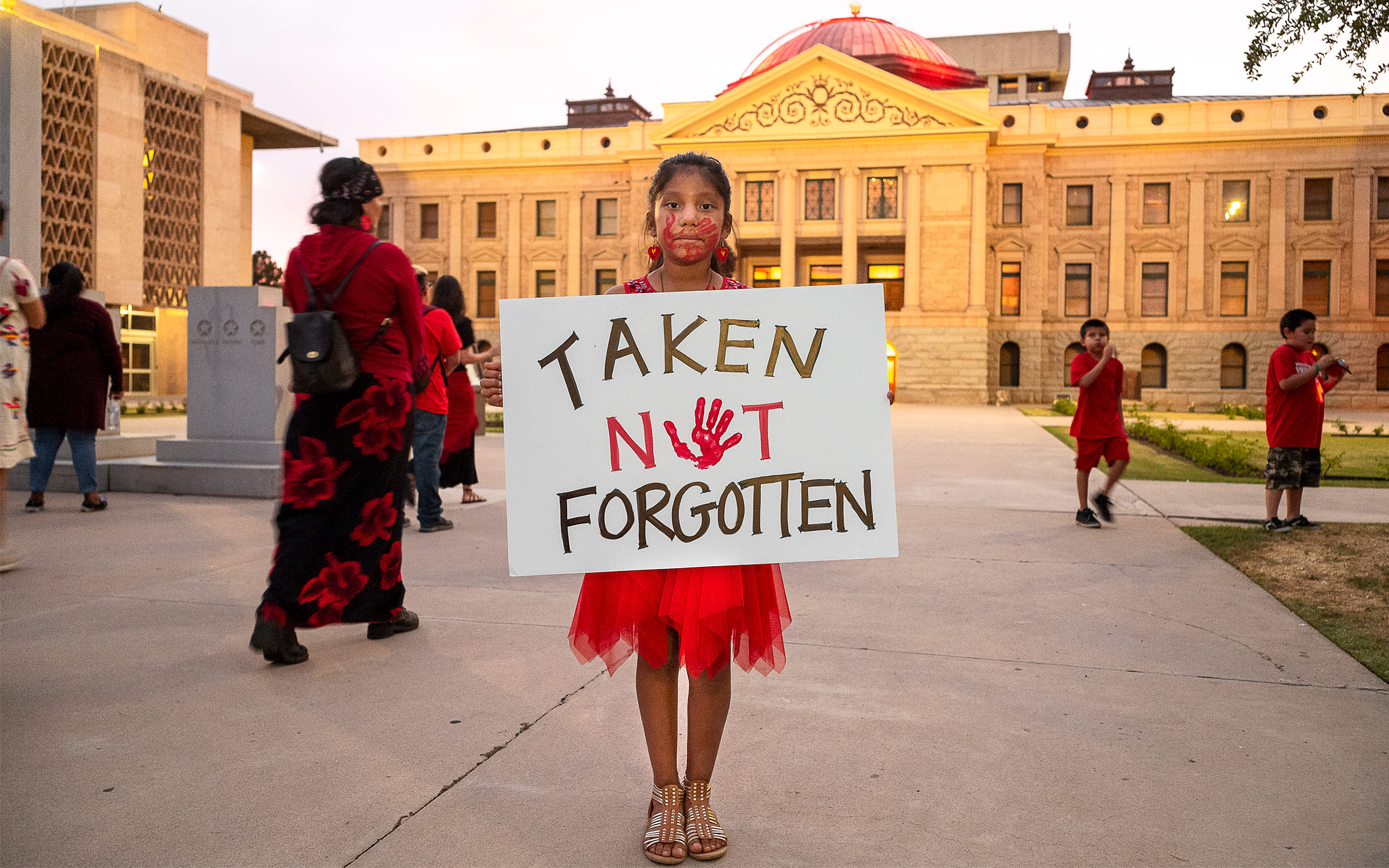 Tyttö pukeutunut punaiseen, pitää kylttiä: "Taken, not forgotten"